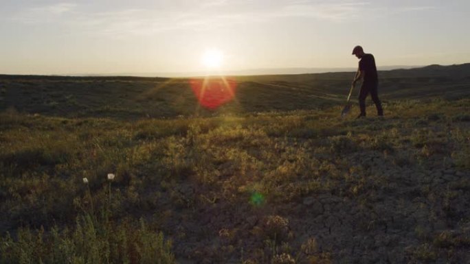 一个三十多岁的白人男子戴着帽子和工作手套，在日落时用铲子在沙漠中挖了一个洞