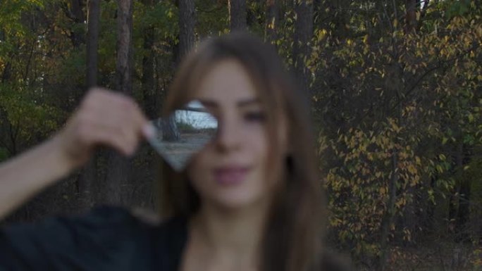 年轻的高加索女孩在她的脸前拿着破碎的镜子碎片。秋天的森林和湖泊在裂缝中反射