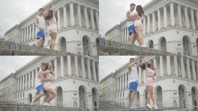 年轻的高加索夫妇在美丽建筑的背景下在城市广场上跳舞。开朗的男人和女人在城市楼梯户外表演。现代艺术概念