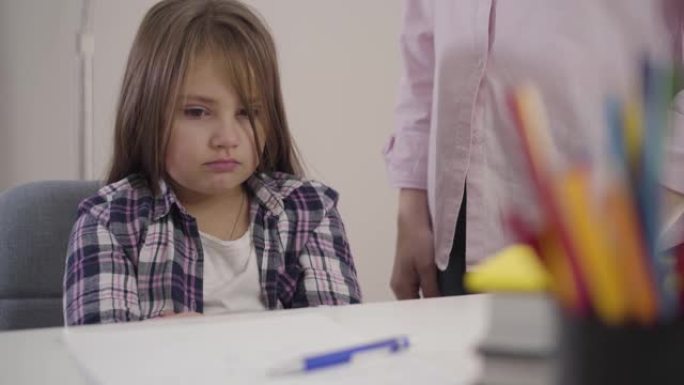 小高加索女学生坐在桌子上的肖像，当她的母亲在她面前摇晃笔时，面部表情不安。筋疲力尽的孩子不想学习。教