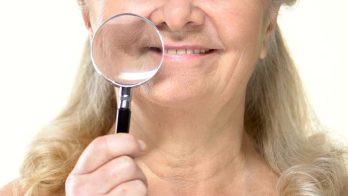 老年女性在皱纹老脸上拿着放大镜，抗衰老化妆品