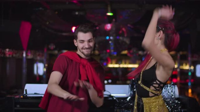 开朗的年轻白人男女在DJ控制器旁边的夜总会跳舞。英俊的男DJ和诱人的女围棋舞者在迪斯科中娱乐。电影院
