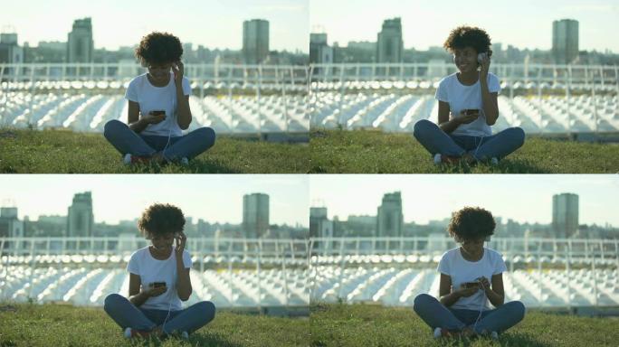 年轻的非洲裔美国妇女戴着耳机听音乐，坐在草坪上，放松