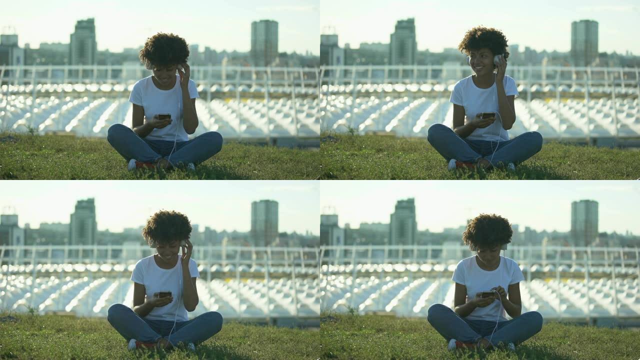 年轻的非洲裔美国妇女戴着耳机听音乐，坐在草坪上，放松
