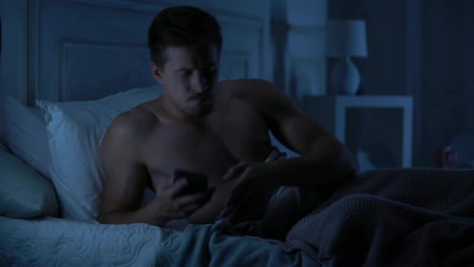 英俊的男性躺在床上，在智能手机上保留令人失望的短信