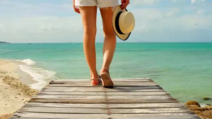 女人戴着海帽她很高兴，她走在木桥上，看着海滩海边，云和蓝天是无尽的背景。