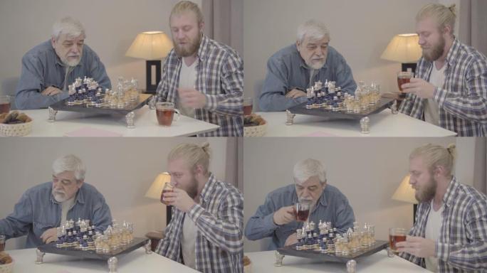 成年的儿子和成熟的父亲在家喝茶作为下棋。快乐的高加索男人在室内一起度过周末的肖像。快乐，爱好，生活方