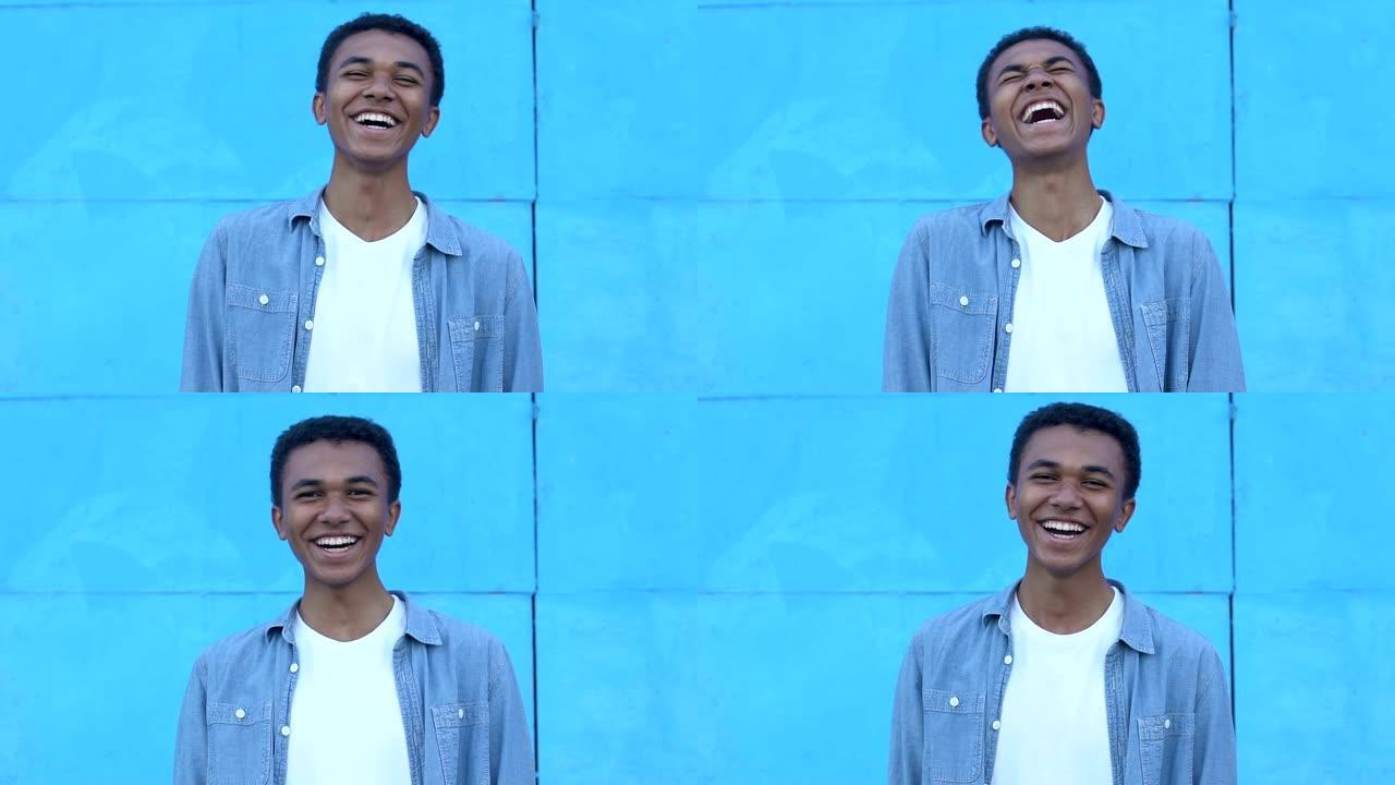 激动的非洲裔美国少年男孩在蓝色背景上大笑，真诚的幸福