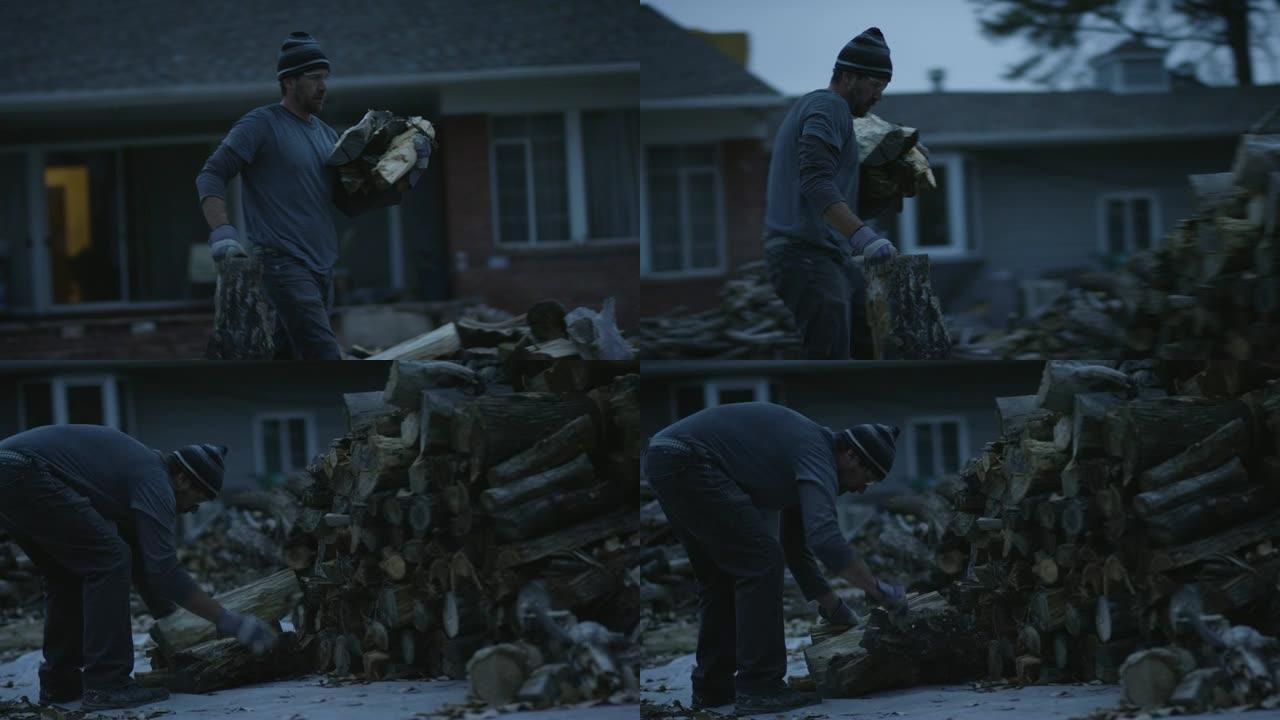 一个四十多岁的白人男子戴着针织帽，安全眼镜和防护手套，在黄昏时将柴火堆在房子旁边的柴堆中。