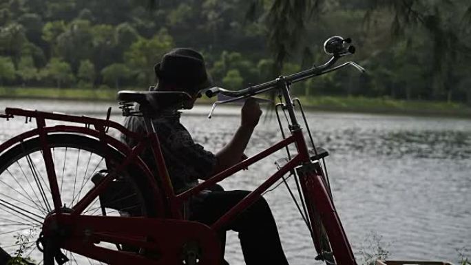 该名男子坐在自行车旁，在湖边扔纸飞机。