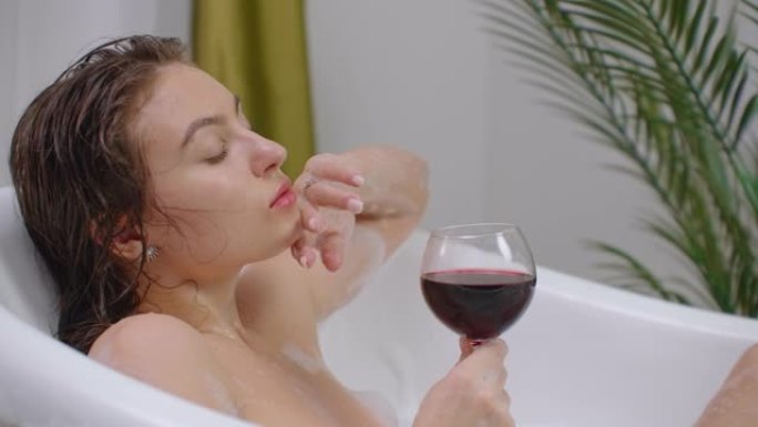 一个诱人的黑发女子躺在泡沫浴中放松和喝红酒。在一个热的泡泡浴中恢复和放松