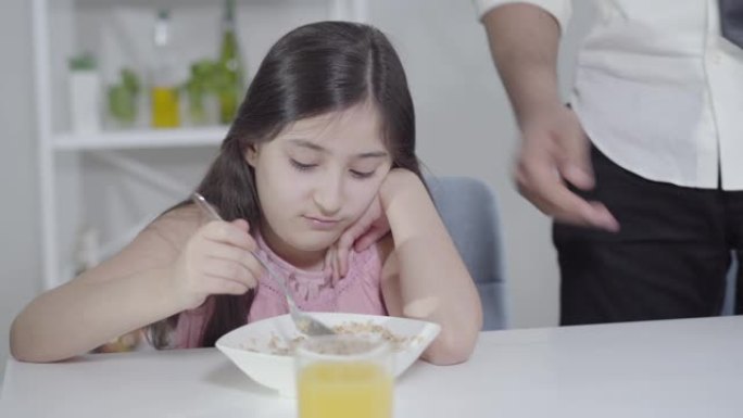 无法辨认的父亲在情感上打手势，并在没有食欲的小女孩在盘子里混合燕麦片时做出迟到的手势。女儿和匆忙的商