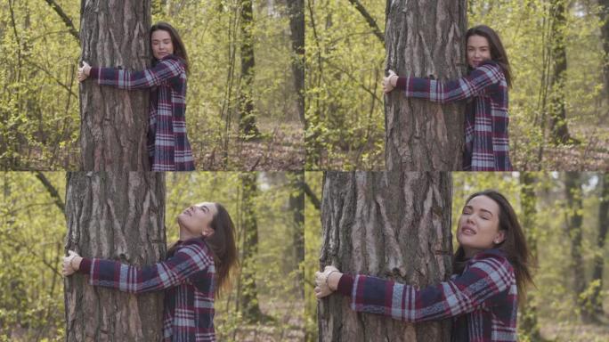 年轻的黑发女人在森林里拥抱树干，微笑着。在阳光明媚的春天或秋天，相机接近迷人的高加索女孩与大自然联系