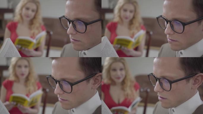 戴着眼镜的年轻疲惫的男人看书，在前景中睡着，而卷曲的金发女人在背景中学习。