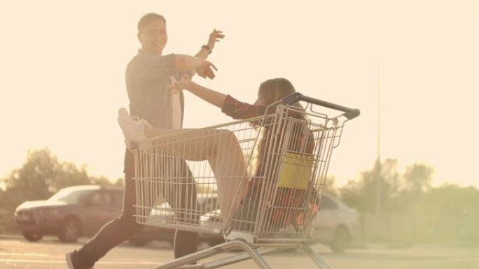 一个年轻的女性和男性在户外购物手推车上玩得开心的侧视图。多种族的年轻人在购物车上比赛。在停车区和他们