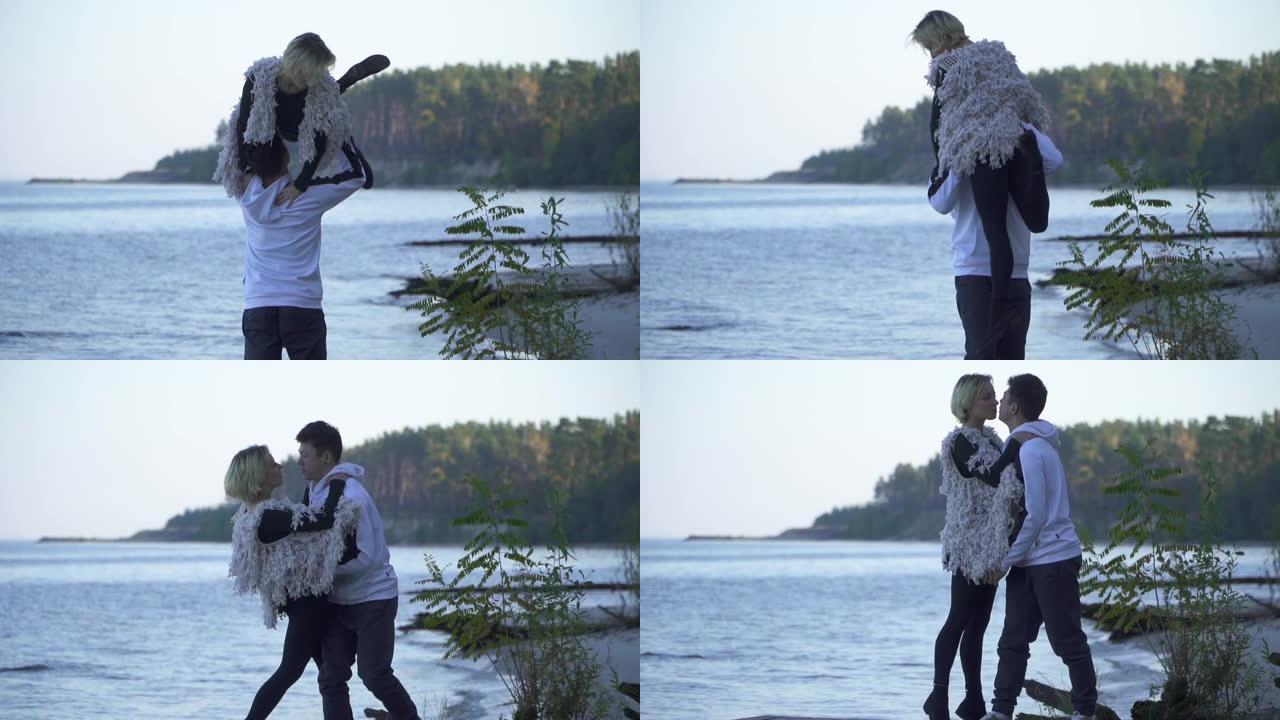 亚洲人在湖泊和森林的背景下将他的白人女友抱在头上。穿着便装的青少年站在沙质河岸上。户外自由式的跨种族