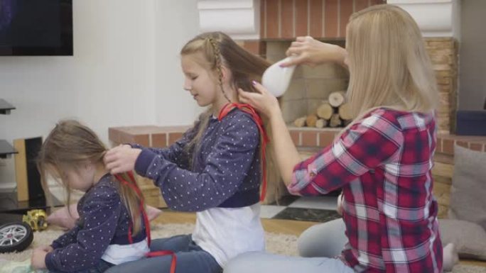 金发碧眼的白人妇女在为妹妹编织辫子时梳理大女儿的头发的侧视图。母亲在室内和孩子一起度过空闲时间。幸福