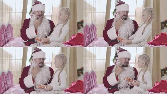 惊讶的高加索女孩看着穿着圣诞老人服装的父亲。假胡子脱掉，男人摸他的脸。爸爸和女儿在平安夜在家玩得开心