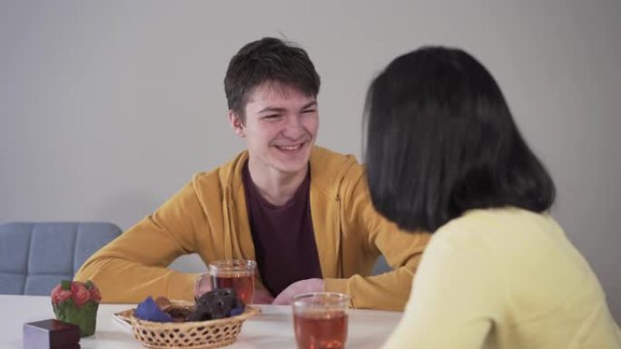微笑的高加索黑发男孩和母亲坐在桌旁喝茶和糖果的肖像。积极的青少年在家和父母一起度过空闲时间。团结、快