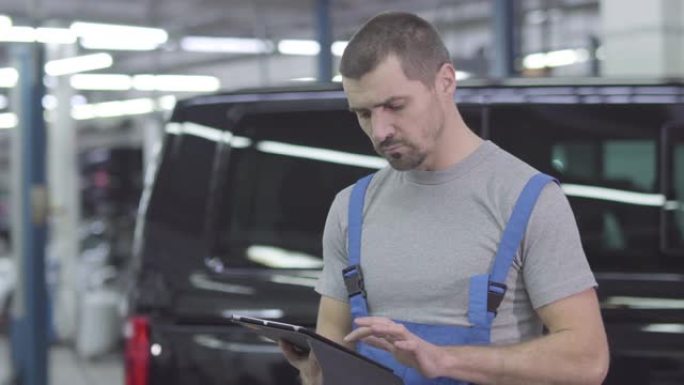 年轻英俊的白人男子穿着工作服站在平板电脑的背景下，黑色汽车。在修理厂工作的严肃的男性汽车修理工。自信