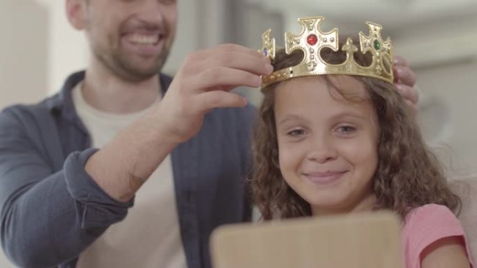 肖像父亲把皇冠戴在照镜子的卷发女孩的头上。