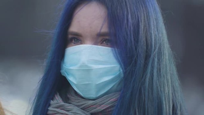 蓝色头发和蓝色眼睛戴着防护面具的年轻女子的特写脸。疫情爆发时站在城市街道上的女人肖像。危险，危险，大