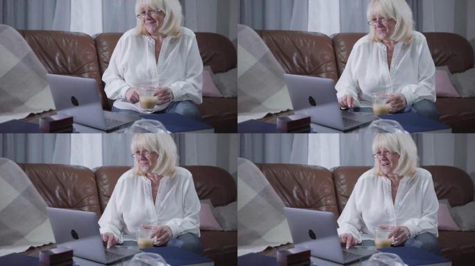 积极的高加索退休人员坐在沙发上，拿着咖啡杯，看着笔记本电脑屏幕。资深金发女子在家上网。消遣、休闲、社