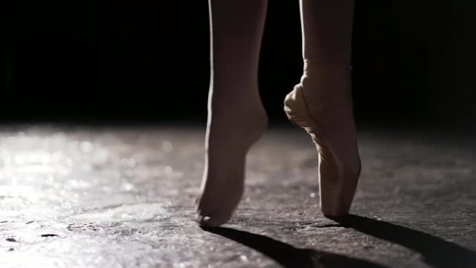 在工作室的黑色背景上，女性纤细的双腿站在聚光灯下。一只脚在足尖，另一只脚没有鞋。特写。