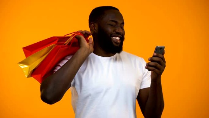 快乐的美国黑人拿着购物袋和智能手机，在网上购物