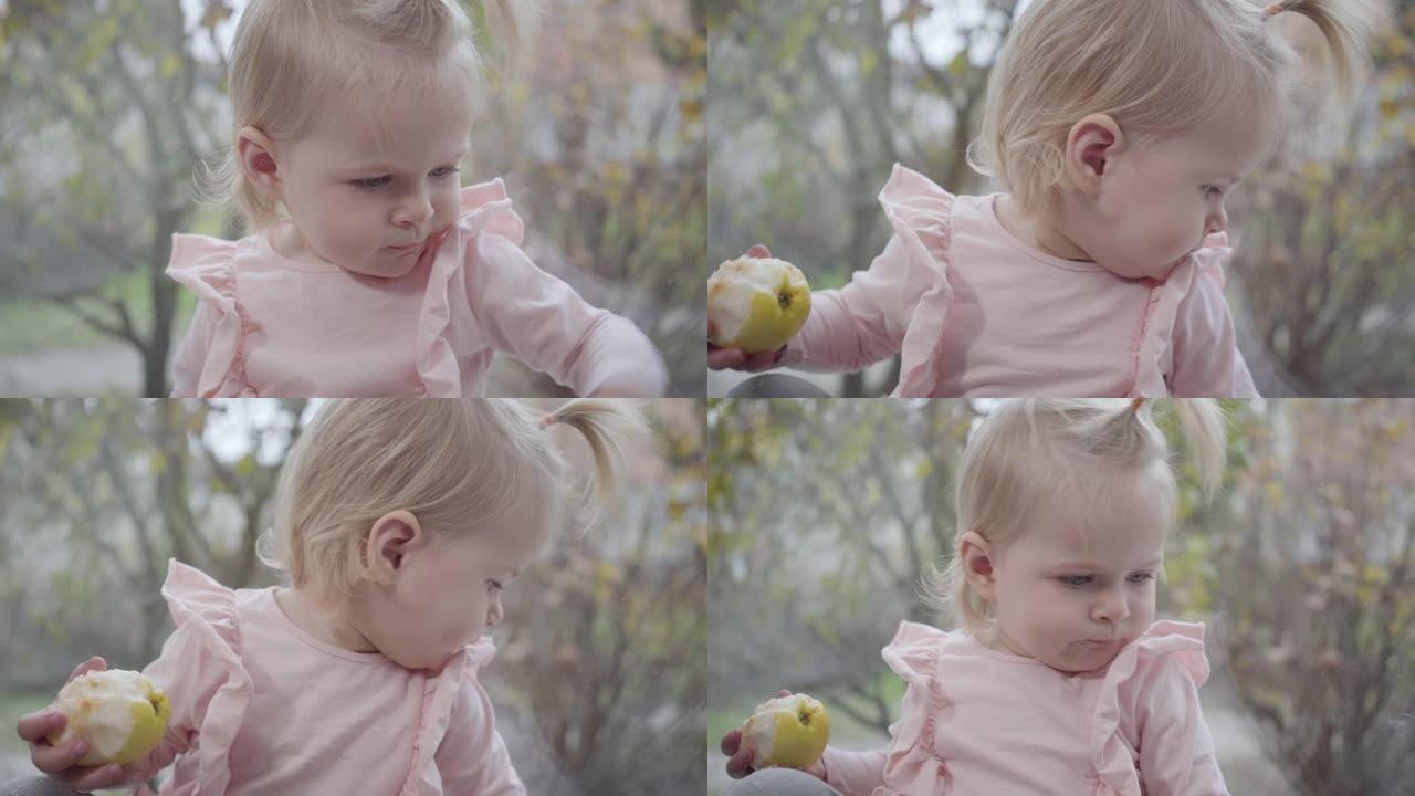可爱的高加索女婴的特写肖像，灰色的眼睛和金色的头发坐在窗台上。穿着粉红色上衣的孩子伸手对着相机吃苹果