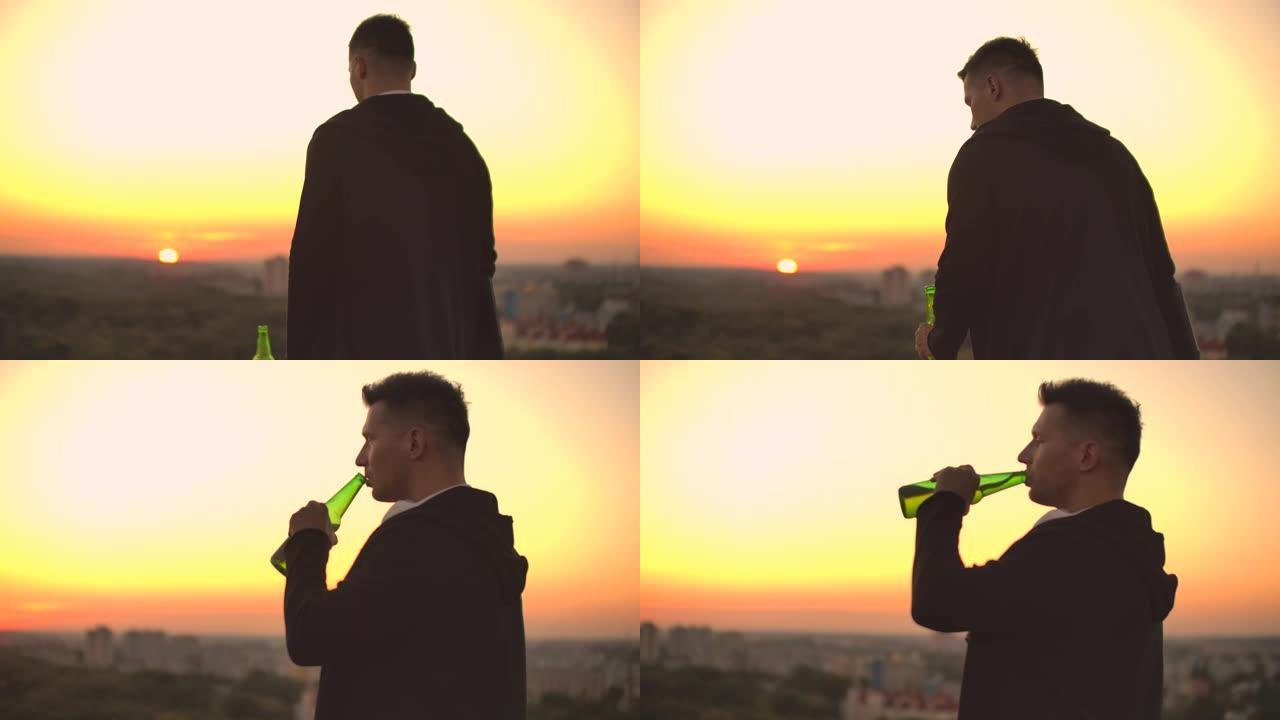 从后面看，一个男人在日落时走在屋顶上，喝啤酒，看着城市的美丽景色