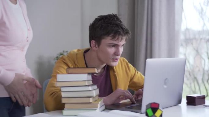 年轻的高加索黑发男孩在笔记本电脑上玩耍，因为他的母亲想出了一堆书。女人把文学摆在桌子上，玩家与父母争