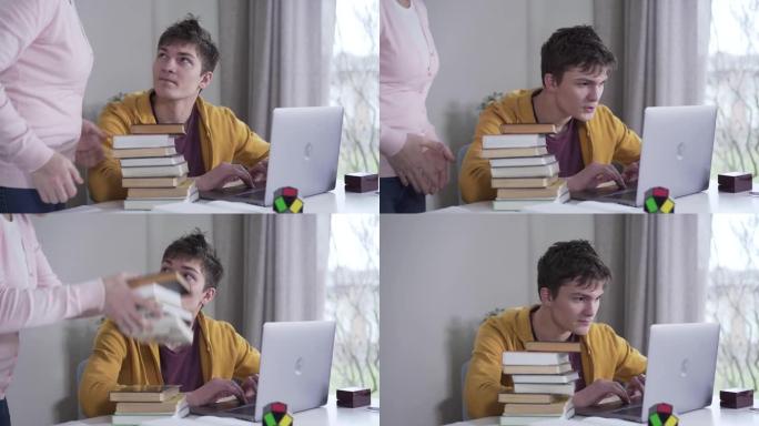 年轻的高加索黑发男孩在笔记本电脑上玩耍，因为他的母亲想出了一堆书。女人把文学摆在桌子上，玩家与父母争