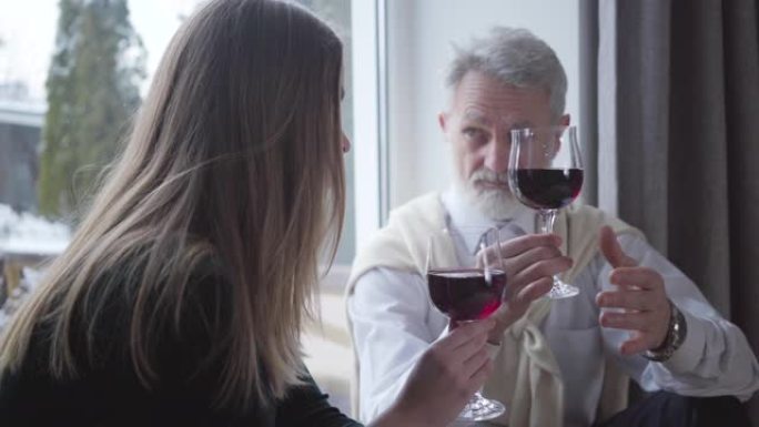 手握酒杯的英俊高级男子在室内与年轻的黑发白人女子交谈。无忧无虑优雅的退休人员与年轻伴侣共度夜晚。年龄