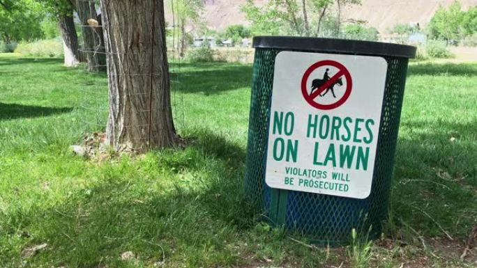 在阳光明媚的日子里，在公园的金属垃圾桶上悬挂着 “草坪上禁止马匹; 违规者将被起诉” 的标志