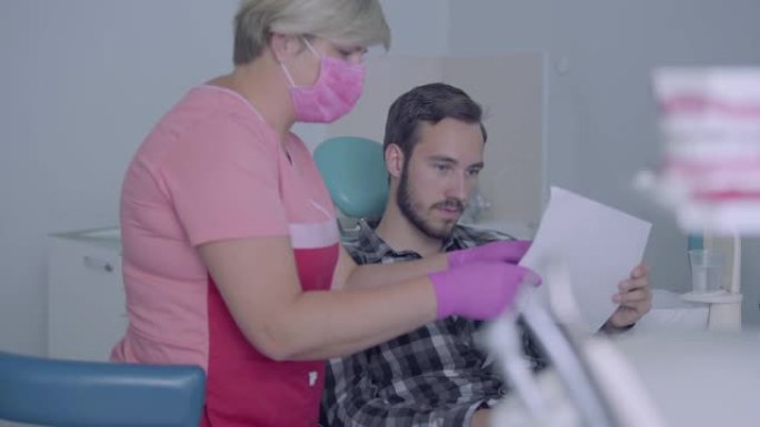 女牙医i医用口罩和手套向男性患者展示他的牙齿图片。