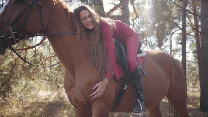 年轻快乐的高加索女性马术者躺在马背上爱抚她的宠物。美丽的棕色马站在秋天的森林里，年轻的骑手在他的背上