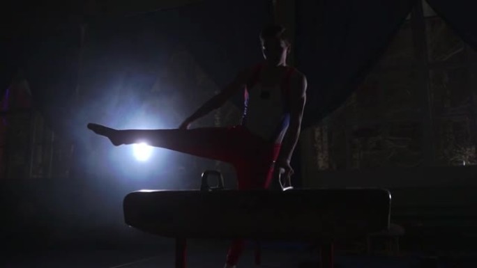 男体操运动员在黑暗的房间里慢动作在烟雾中进行鞍马运动。体育促进健康和成就