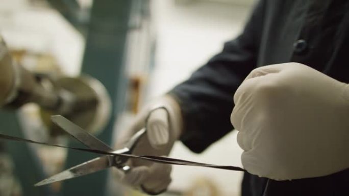 一名戴着防护手套的技术人员在室内制造设施中使用一把金属剪刀切割一串黑色碳纤维