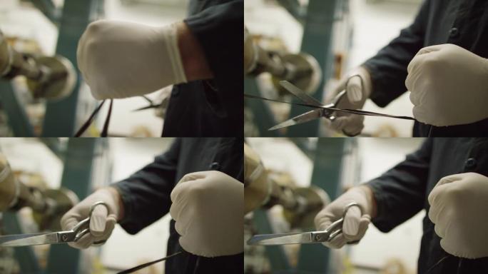 一名戴着防护手套的技术人员在室内制造设施中使用一把金属剪刀切割一串黑色碳纤维