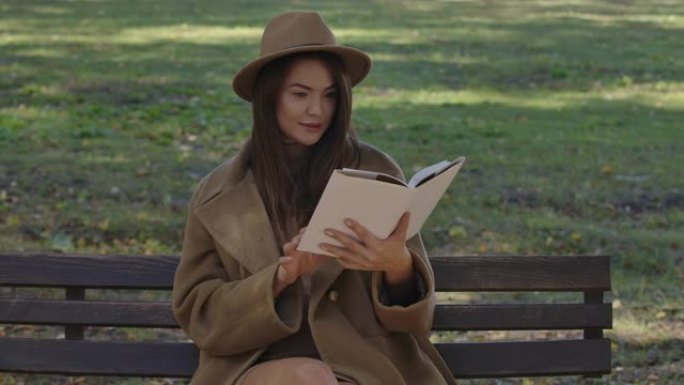 可爱的白人女学生，穿着优雅的棕色衣服，坐在长凳上翻书。迷人聪明的欧洲女人在秋季公园读书。电影院4k镜