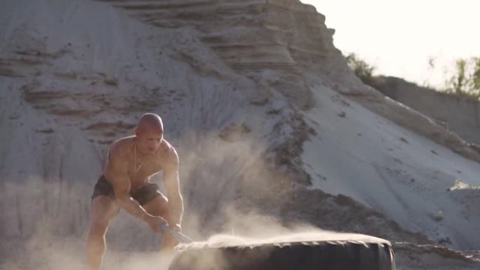 秃头强人在沙山的一个巨大轮子上慢动作击中锤子。摔跤运动员的力量和耐力训练。