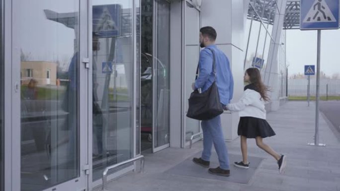 穿着休闲蓝色衣服的成年白人男子和长卷发的小女孩进入机场大门。父亲和女儿一起旅行。幸福的家庭周末出国。