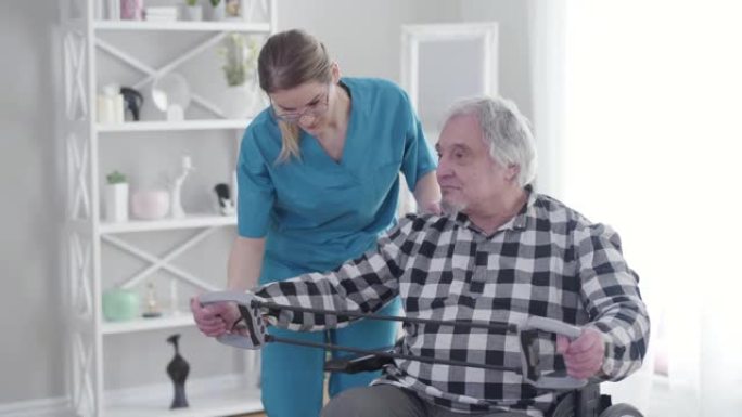 穿着蓝色制服和眼镜的专业护士帮助老年残疾人在疗养院锻炼。拉伸松紧带时支持退休人员的无效投标。物理治疗