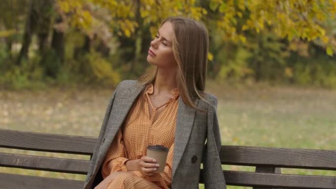 美丽的高加索女人坐在秋天公园的长椅上喝杯咖啡。迷人的女孩摇着棕色的长发，把目光移开。电影院4k镜头P
