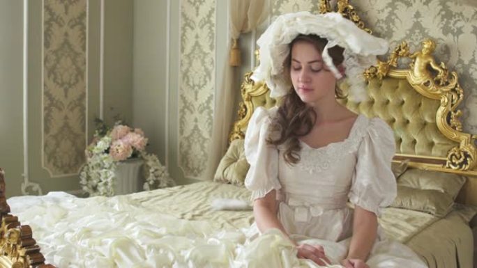 穿着白色复古舞会礼服和帽子的年轻美女坐在巨大的金色床上，用手机隐藏