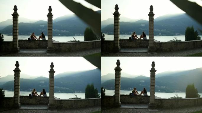 两名年轻妇女坐在石墙上，被柱子框住，俯瞰湖泊和山脉