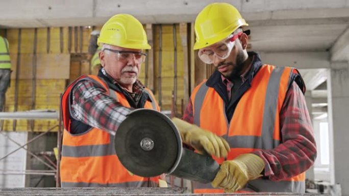 高级男性建筑工人向年轻的男学徒展示如何握住大型角磨机切穿钢网