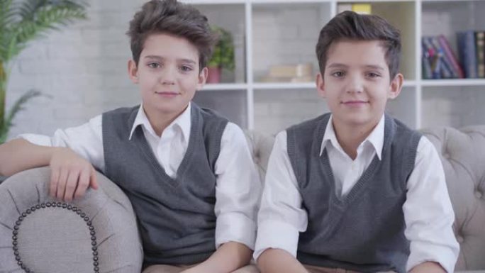 同卵双胞胎兄弟互相看着对方，转向镜头并微笑的肖像。优雅的白人儿童穿着类似的衣服坐在室内的沙发上。家庭