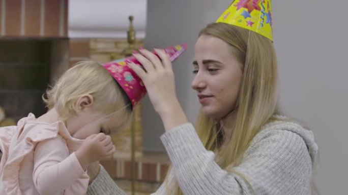 年轻的白人妇女把党帽戴在小女孩的头上。穿着粉色上衣的漂亮宝贝和妈妈一起庆祝她的第一个生日。电影院4k